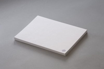 A stack of plain white paper sheets lies against a grey background. The edges of the papers are visible, and in the bottom corner of the top sheet, there is a small circular logo in blue ink.