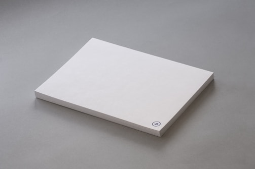 A stack of plain white paper sheets lies against a grey background. The edges of the papers are visible, and in the bottom corner of the top sheet, there is a small circular logo in blue ink.