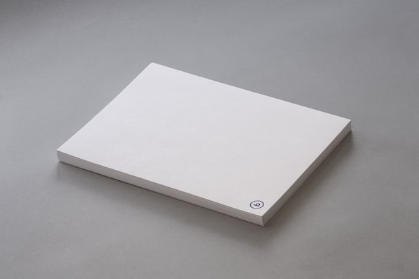 A stack of plain white paper sheets lies against a grey background. The edges of the papers are visible, and in the bottom corner of the top sheet, there is a small circular logo in blue ink.