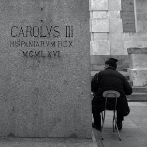 grayscale photography of man sitting beside a concrete wall