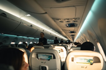 The interior of an airplane cabin is depicted, revealing rows of occupied seats with passengers seated. Overhead compartments are visible above the seating area. A flight attendant is standing in the aisle attending to passengers. The lighting suggests daytime, with natural light streaming through the windows and soft artificial lighting illuminating the cabin.