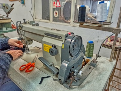 A sewing machine sits on a table with a pair of hands working on fabric. The machine is equipped with several spools of thread, including black, green, and white. An orange pair of scissors rests on the table beside the machine. The setting appears to be a small, organized sewing workshop.