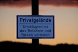 A sign with German text indicates private property, prohibiting unauthorized access and parking. It is displayed against a backdrop of a clear sky during sunset, with a gradient from orange to a darker hue near the horizon.