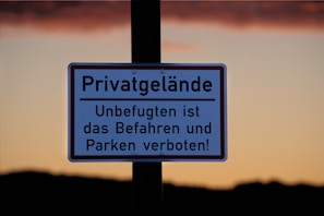 A sign with German text indicates private property, prohibiting unauthorized access and parking. It is displayed against a backdrop of a clear sky during sunset, with a gradient from orange to a darker hue near the horizon.