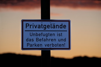 A sign with German text indicates private property, prohibiting unauthorized access and parking. It is displayed against a backdrop of a clear sky during sunset, with a gradient from orange to a darker hue near the horizon.