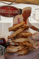 A vendor with white hair and a white hat, wearing a white shirt and orange vest, is arranging a stack of freshly baked, large ring-shaped bread on a stall. There is a sign above him with the text 'AMBELLERIA ALONZI'. The atmosphere is that of a marketplace or street fair.