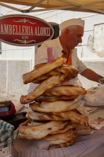 A vendor with white hair and a white hat, wearing a white shirt and orange vest, is arranging a stack of freshly baked, large ring-shaped bread on a stall. There is a sign above him with the text 'AMBELLERIA ALONZI'. The atmosphere is that of a marketplace or street fair.