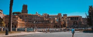A historic architectural site features ancient Roman ruins with multiple arched structures and stone buildings. The scene is lively with people walking around the area, and a clear blue sky overhead adds to the vibrant atmosphere.