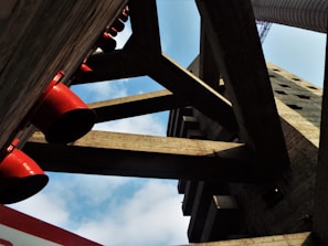 A perspective view of an architectural structure with large concrete beams and cylindrical red pipes. The arrangement of the beams creates geometric patterns against a backdrop of a partly cloudy blue sky.