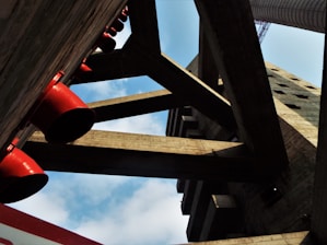 A perspective view of an architectural structure with large concrete beams and cylindrical red pipes. The arrangement of the beams creates geometric patterns against a backdrop of a partly cloudy blue sky.