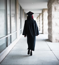 A person wearing a graduation gown and cap is walking down a corridor. The corridor is lined with large windows on one side and stone pillars on the other.