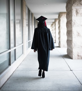A person wearing a graduation gown and cap is walking down a corridor. The corridor is lined with large windows on one side and stone pillars on the other.