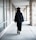 A person wearing a graduation gown and cap is walking down a corridor. The corridor is lined with large windows on one side and stone pillars on the other.