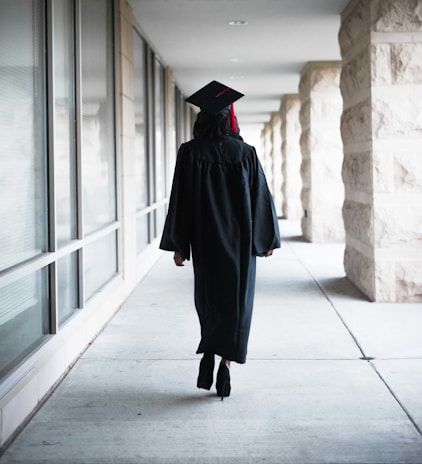A person wearing a graduation gown and cap is walking down a corridor. The corridor is lined with large windows on one side and stone pillars on the other.