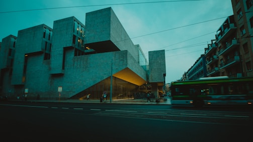 A modern architectural building with sharp, angular lines and a concrete facade dominates the scene. It is situated on a city street with a blurred motion of a green bus passing by. The building's design includes protruding sections and large windows, illuminated from within, casting a warm glow.