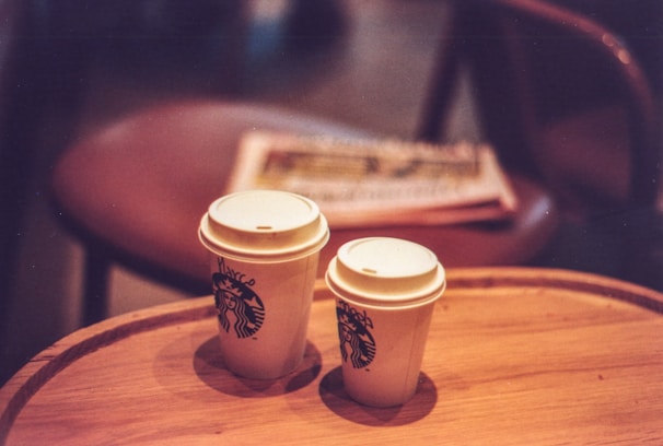 Two disposable coffee cups with lids are placed on a wooden table. Both cups feature the Starbucks logo, with one cup having handwriting on it. In the background, there is a blurred chair and a stack of newspapers, creating a cozy coffee shop ambiance.