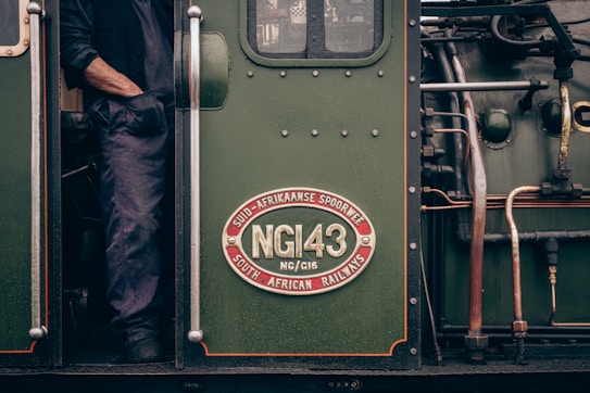 A detailed view of a vintage locomotive, showcasing a metallic plaque with 'NG143' and 'South African Railways' in both English and Afrikaans. A person is partially visible with one arm resting against the door frame, dressed in dark blue work attire. The locomotive is painted in a deep green tone with various mechanical components, including pipes, visible on the side.