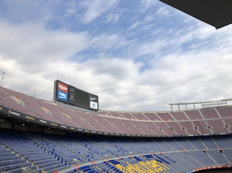 An empty stadium with multiple tiers of seating is shown. The seats are colored blue, yellow, and red, indicating various sections. A large electronic scoreboard rests at the top with advertisements for companies including Rakuten, Beko, Nike, and Caixa Bank. The sky above is partly cloudy with patches of blue visible.