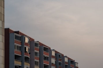 A row of residential apartment buildings with multiple floors is seen against a cloudy sky. The buildings have a modern architectural design with a combination of red bricks and concrete, and glass windows with balconies.