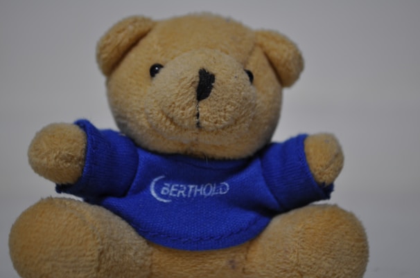 A plush teddy bear wearing a light blue scarf sitting on a white blanket.