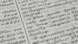 Close-up of a page with Tamil script written in neat black letters, showing two columns of text. The focus is on the middle area, with surrounding text slightly blurred.