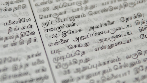 Close-up of a page with Tamil script written in neat black letters, showing two columns of text. The focus is on the middle area, with surrounding text slightly blurred.