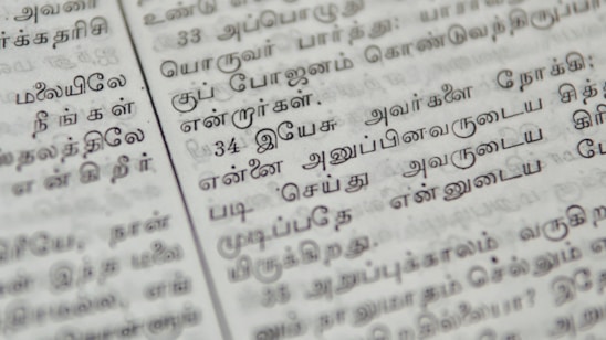 Close-up of a page with Tamil script written in neat black letters, showing two columns of text. The focus is on the middle area, with surrounding text slightly blurred.