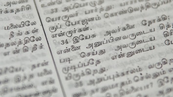 Close-up of a page with Tamil script written in neat black letters, showing two columns of text. The focus is on the middle area, with surrounding text slightly blurred.