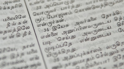 Close-up of a page with Tamil script written in neat black letters, showing two columns of text. The focus is on the middle area, with surrounding text slightly blurred.
