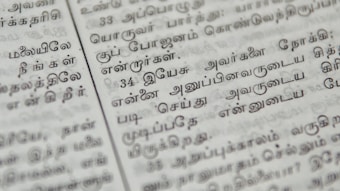 Close-up of a page with Tamil script written in neat black letters, showing two columns of text. The focus is on the middle area, with surrounding text slightly blurred.
