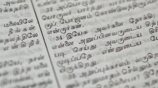 Close-up of a page with Tamil script written in neat black letters, showing two columns of text. The focus is on the middle area, with surrounding text slightly blurred.