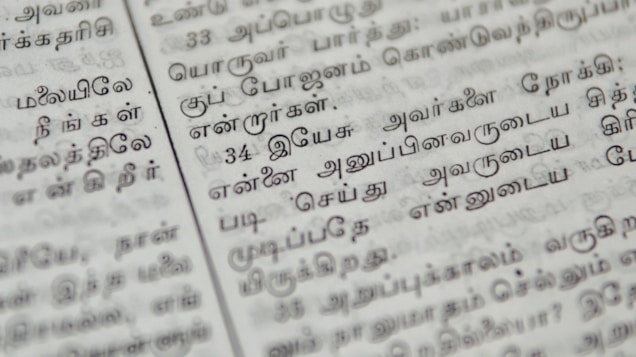 Close-up of a page with Tamil script written in neat black letters, showing two columns of text. The focus is on the middle area, with surrounding text slightly blurred.