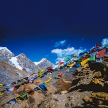 Colorful prayer flags flutter in the wind against a backdrop of majestic snow-capped mountains and a clear blue sky. The rugged terrain is filled with rocky slopes and peaks, creating a striking contrast with the vivid fabric of the flags.