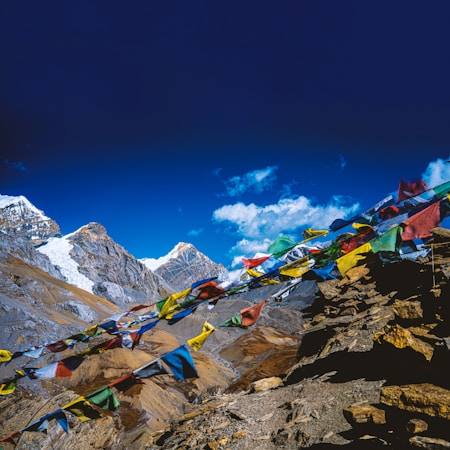 Colorful prayer flags flutter in the wind against a backdrop of majestic snow-capped mountains and a clear blue sky. The rugged terrain is filled with rocky slopes and peaks, creating a striking contrast with the vivid fabric of the flags.