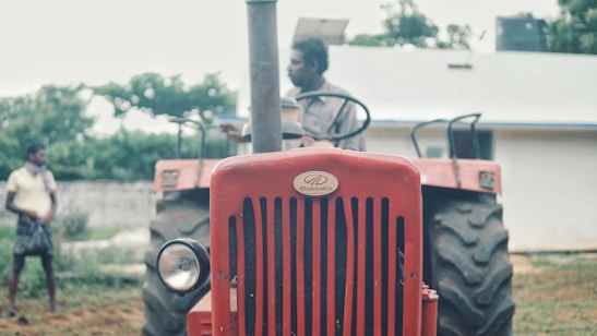 A red tractor with a focus on its front grille and headlight. A person is seated on the tractor while another person stands in the background. The surrounding environment includes trees and a white building.