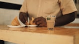 A person in a beige shirt is seated at a wooden table, preparing to eat samosas placed on small white plates. In the foreground, there's a small glass of chai.