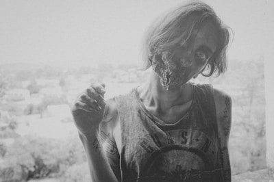 A horror-themed black and white image depicting a person with a gruesome facial injury or special effects makeup. The person's hair is disheveled, and they wear a sleeveless shirt with tattoos visible on the arms. The background appears to be an urban setting out of focus.
