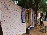 A collection of vibrant, intricately designed tapestries and fabrics hang outdoors among trees. The artwork features various colors and patterns, including spirals and abstract figures. The setting appears natural and serene, with dirt ground and greenery surrounding the area.