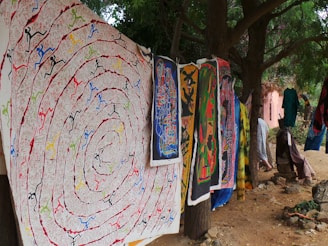 A collection of vibrant, intricately designed tapestries and fabrics hang outdoors among trees. The artwork features various colors and patterns, including spirals and abstract figures. The setting appears natural and serene, with dirt ground and greenery surrounding the area.