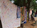 A collection of vibrant, intricately designed tapestries and fabrics hang outdoors among trees. The artwork features various colors and patterns, including spirals and abstract figures. The setting appears natural and serene, with dirt ground and greenery surrounding the area.