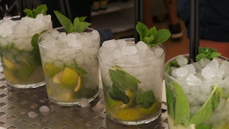 Four mojito cocktails are arranged in a row on a metallic surface, each glass filled with crushed ice, lime wedges, and fresh mint leaves, topped with a black straw.