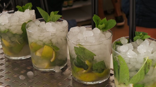 Four mojito cocktails are arranged in a row on a metallic surface, each glass filled with crushed ice, lime wedges, and fresh mint leaves, topped with a black straw.