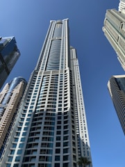 Skyscrapers with modern architectural designs rise against a clear blue sky. The tall buildings feature glass facades and varied geometric patterns, capturing the sense of urban development and soaring heights.