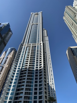 Skyscrapers with modern architectural designs rise against a clear blue sky. The tall buildings feature glass facades and varied geometric patterns, capturing the sense of urban development and soaring heights.