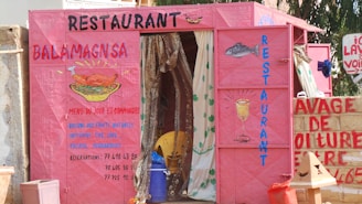 A colorful, makeshift restaurant entrance painted in bright pink with handwritten menus and illustrations of food items. The front displays a fish and a drink, while curtains hang behind the entrance.