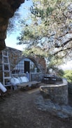A rustic stone patio area with a stone wall and archway is surrounded by lush greenery. There is a wooden ladder leaning against the wall and a cozy seating area with cushions and a low table to the left. To the right, a dining table with chairs is set up, partially shaded by an overhead tree.