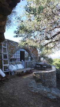 A rustic stone patio area with a stone wall and archway is surrounded by lush greenery. There is a wooden ladder leaning against the wall and a cozy seating area with cushions and a low table to the left. To the right, a dining table with chairs is set up, partially shaded by an overhead tree.