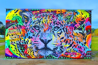 A vibrant and colorful graffiti artwork features a large, detailed depiction of a tiger's face. The mural is covered with a mix of bright colors including pinks, yellows, blues, and greens, interspersed with various graphic elements and text fragments. The eyes of the tiger, painted in gray and black, are prominently centered and draw attention amidst the chaotic background.
