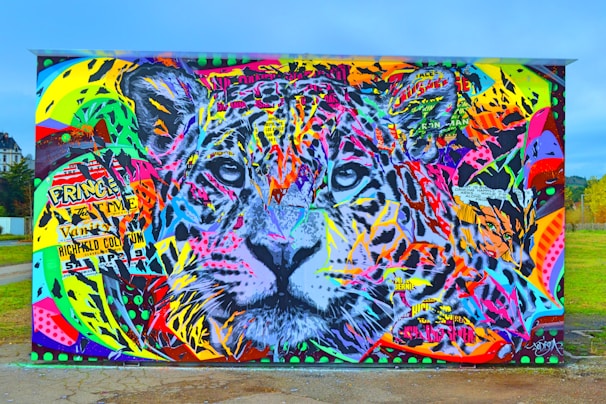 A vibrant and colorful graffiti artwork features a large, detailed depiction of a tiger's face. The mural is covered with a mix of bright colors including pinks, yellows, blues, and greens, interspersed with various graphic elements and text fragments. The eyes of the tiger, painted in gray and black, are prominently centered and draw attention amidst the chaotic background.