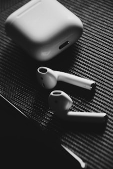 A pair of wireless earbuds and their charging case are placed on a textured surface. The image is in black and white, highlighting the sleek design of the earbuds and the simplicity of the case.